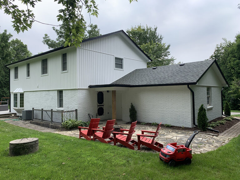 Two-story white house with black roof in Markham, red Adirondack chairs on a stone patio, toy car on the grass, and trees in the background.