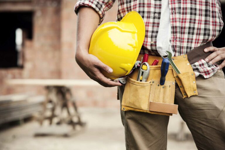 A construction worker in Markham, wearing a tool belt, holds a yellow hard hat while standing at a work site.