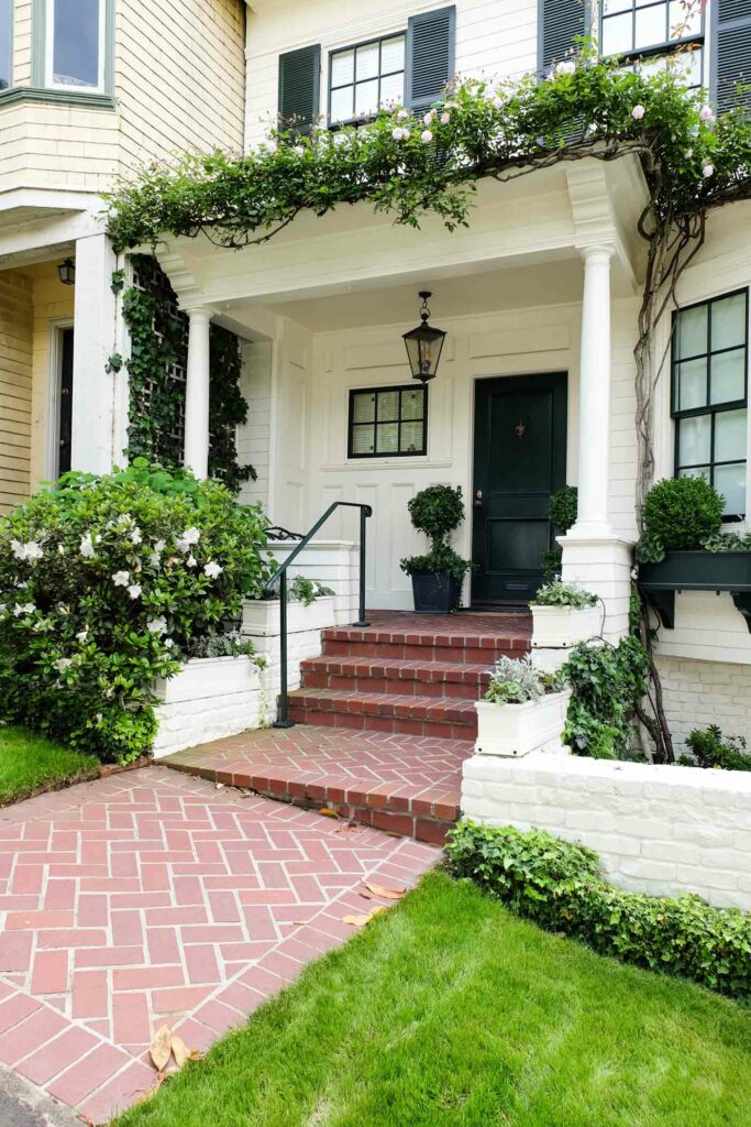 Brick walkway leads to a front porch with white columns, green door and shutters, potted plants, and climbing vines on a charming Markham white house.