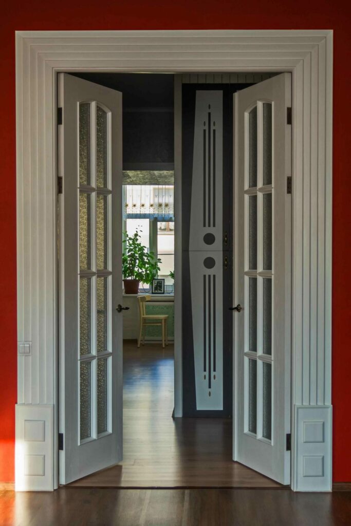 White double doors open to a hallway with a geometric patterned wall, leading to a sunlit Markham room with a window, potted plant, and a chair.