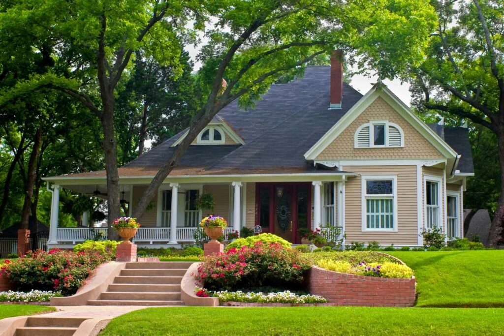 A yellow house with a large front porch in Markham, surrounded by trees, flowers, a manicured lawn, and brick planters along a walkway with steps.