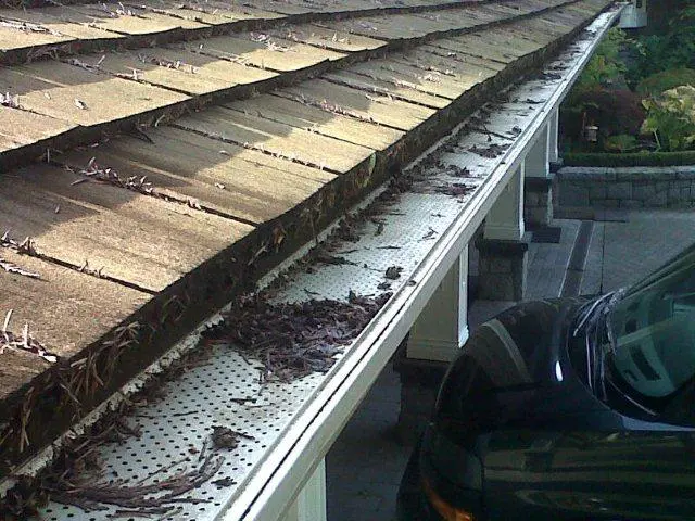 A close-up of a house roof in Markham with leaf guard gutters partially covered by leaves and debris, adjacent to a parked car.