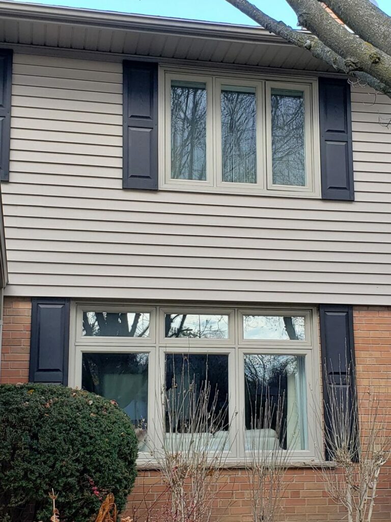 A two-story Markham house with beige siding and brick on the lower level, featuring four black-shuttered windows and visible tree branches overhead.