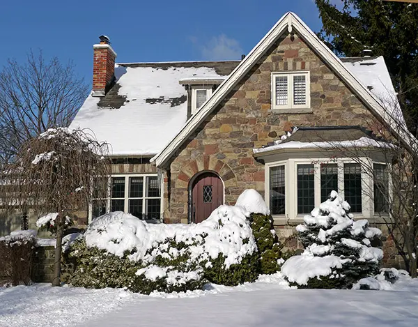 A stone house with a steep roof and a red door sits in Markham, surrounded by snow-covered bushes and trees on a clear winter day.